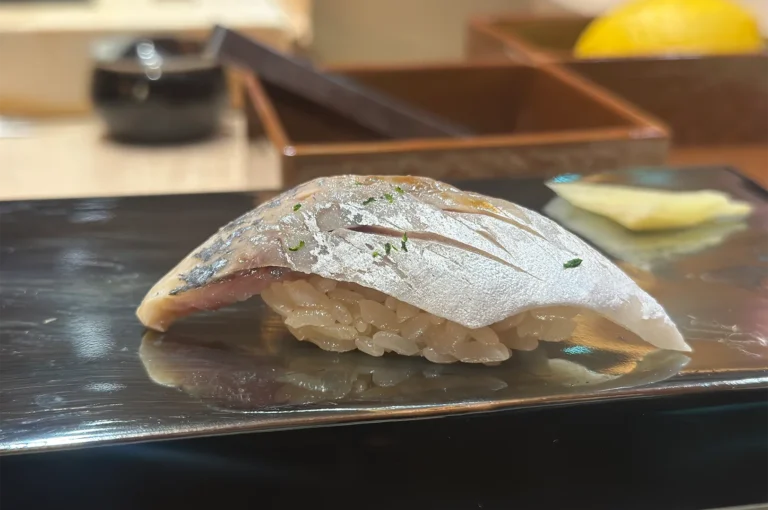 A piece of premium Aji (Spanish Mackerel) nigiri sushi with delicate garnishes served on a black stone plate.