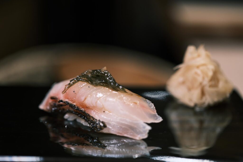 Close-up of a delicate white fish nigiri topped with seasoned kelp on a black lacquer plate at a high-end sushi restaurant.