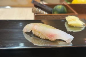 A piece of Madai (Red Sea Bream) nigiri sushi served on a black lacquer plate with pickled ginger in the background.