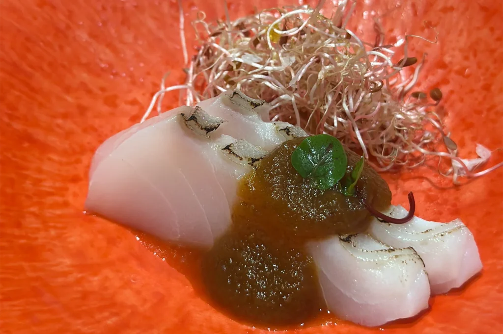 Slices of seared white fish sashimi topped with savory miso glaze and microgreens in a vibrant orange bowl.