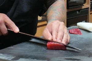 A professional sushi chef with a forearm tattoo carefully slicing a fresh piece of red tuna sashimi with a traditional Japanese knife.