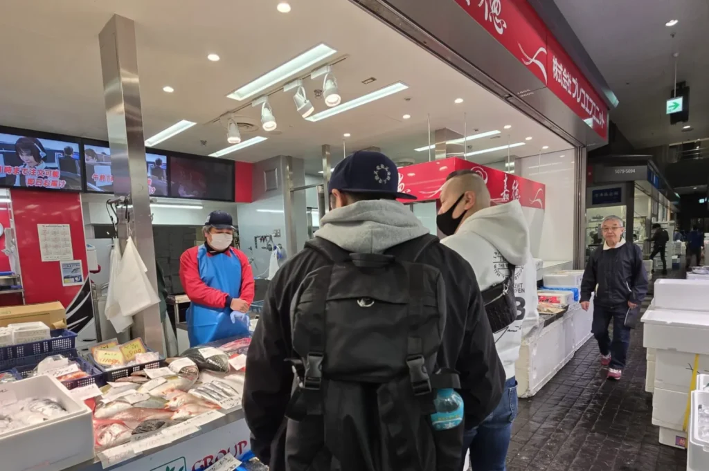Chef Masa browse seafood at a bright fish market stall. The vendor is wearing a mask and gloves. Displayed fish are fresh, and neatly arranged on ice.