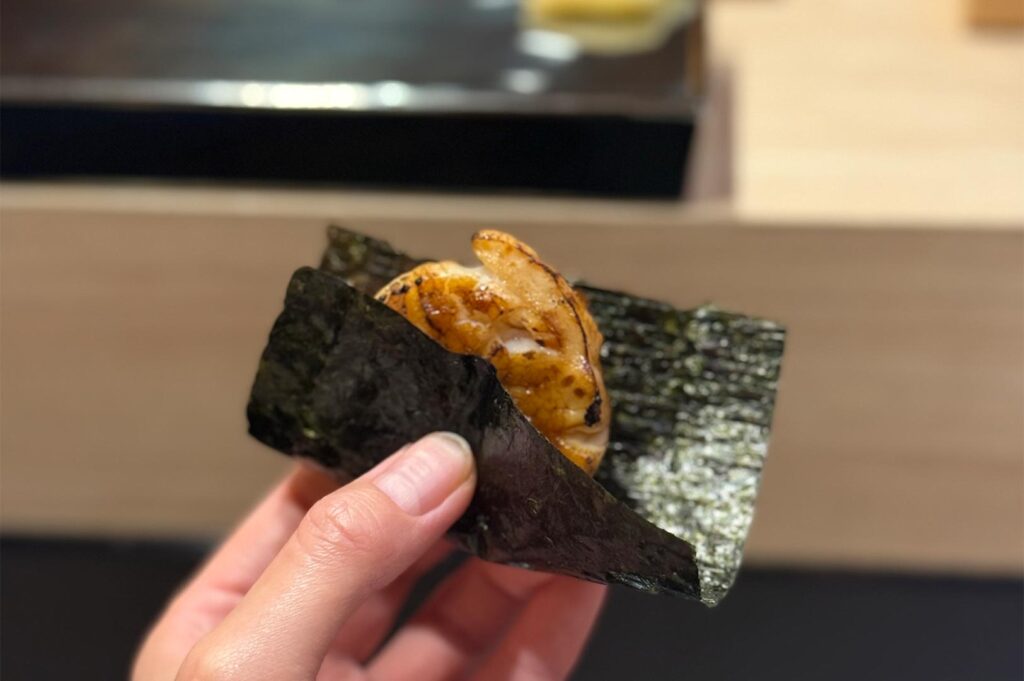 A hand holding a piece of seared aburi scallop wrapped in crispy nori seaweed, showcasing a charred glaze.