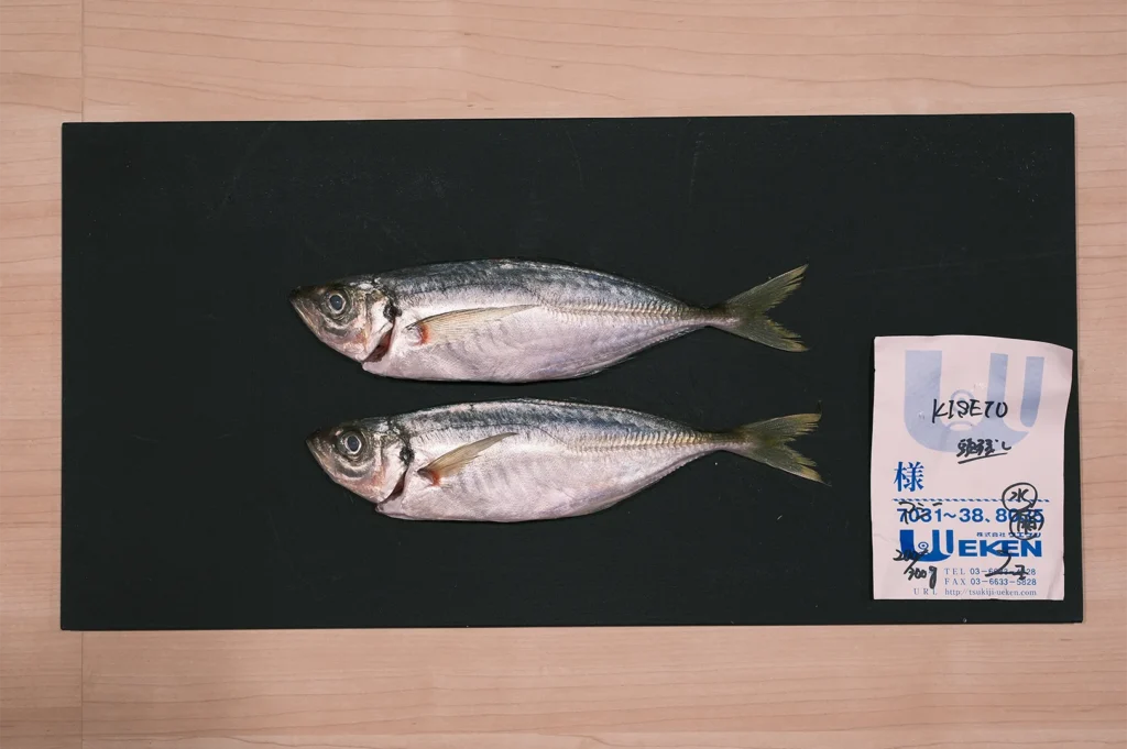 Two fresh whole Japanese horse mackerel (Aji) laid on a black cutting board with a Tsukiji market fish monger receipt.