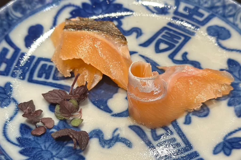 Two delicate pieces of fresh salmon sashimi with skin-on detail, served on a traditional Japanese blue and white patterned plate.