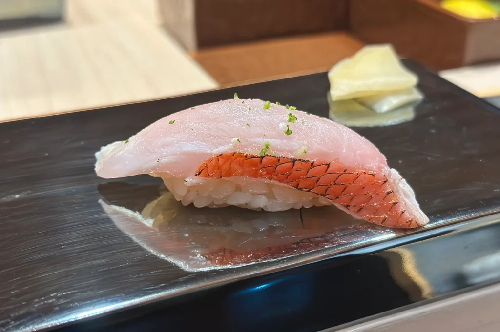 A piece of Kinmedai nigiri sushi featuring a delicate pink fish slice with a distinctive red skin edge, garnished with lime zest on a lacquer plate.