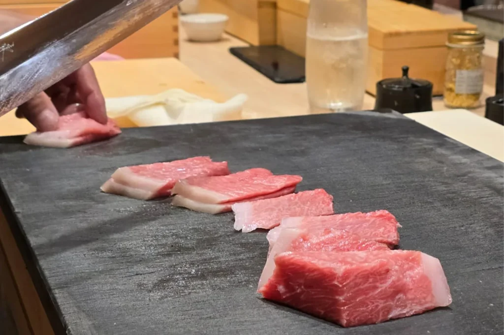 Close-up of premium marbled otoro (fatty bluefin tuna) slices being prepared on a dark slate cutting board at a sushi bar.