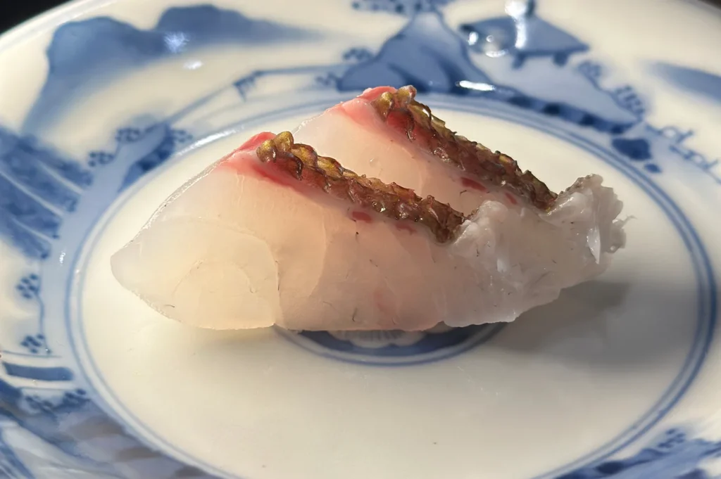 Two slices of fresh Tai snapper sashimi with skin lightly seared, served on a traditional Japanese blue and white ceramic plate.