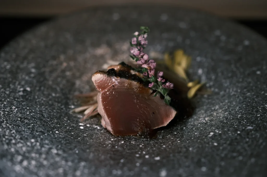 A gourmet slice of seared tuna sashimi served on a textured grey stone plate, garnished with delicate purple shiso flowers.