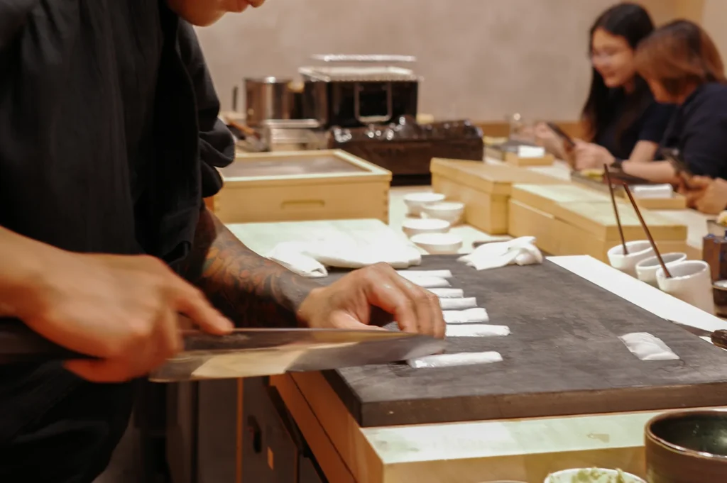 A close-up of a professional sushi chef with arm tattoos skillfully slicing fresh white fish at a wooden omakase counter while diners look on.