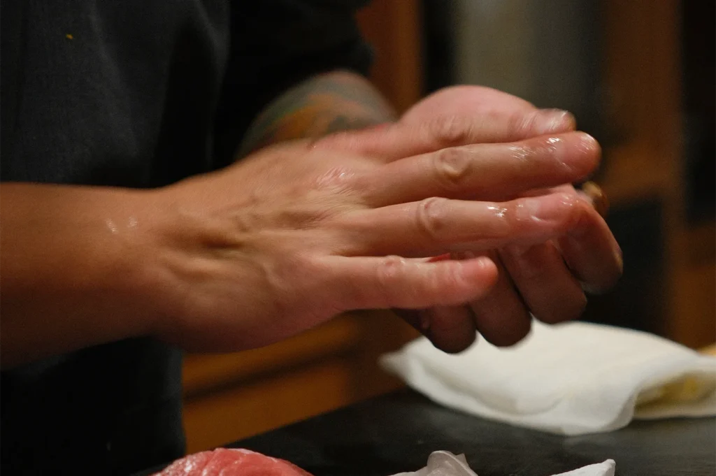A close-up shot of a sushi chef's hands skillfully pressing and shaping a piece of nigiri sushi over a wooden workspace.