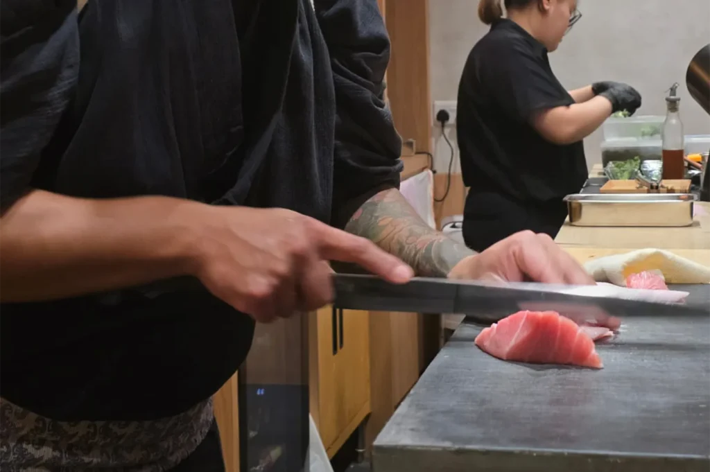 A close-up of a sushi chef’s hands using a traditional Japanese knife to precisely slice a piece of fatty bluefin tuna (otoro) on a stone workspace.