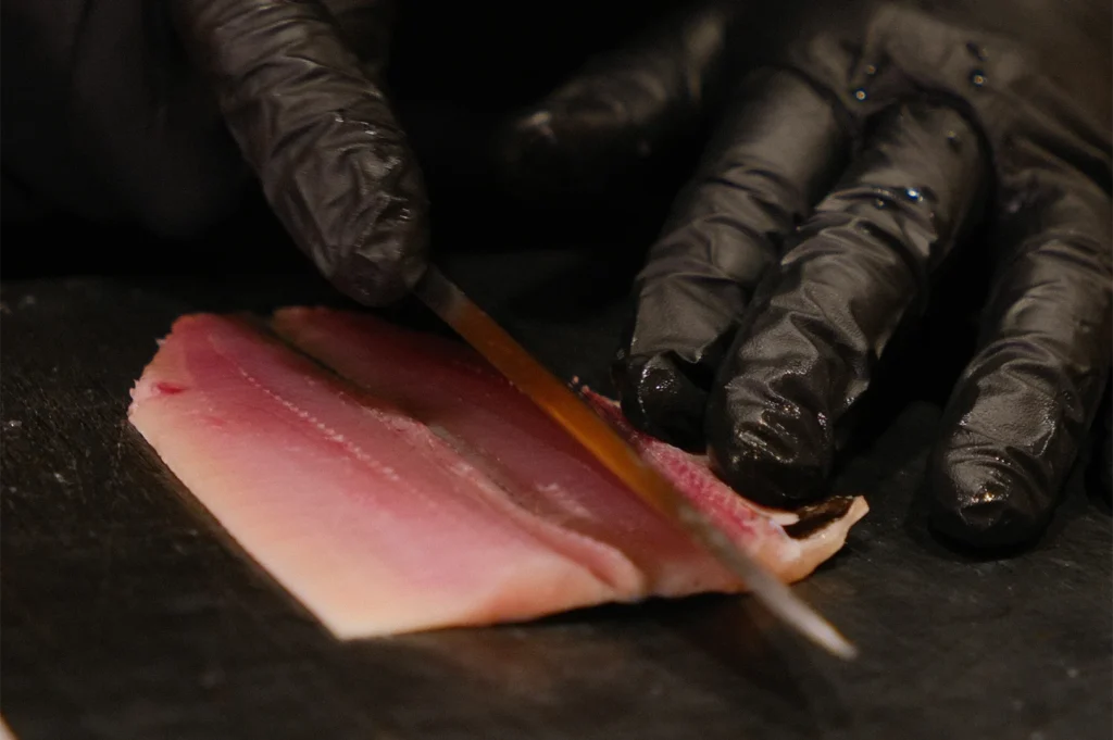 Close-up of a sushi chef in black gloves precisely slicing a fresh pink mackerel fish fillet on a dark cutting board.