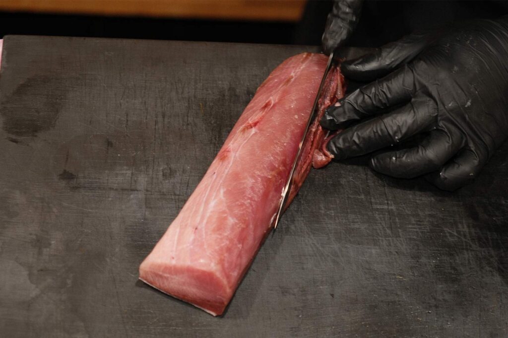 High-angle shot of a sushi chef in black gloves using a traditional yanagiba knife to slice a fresh tuna loin on a dark cutting board.