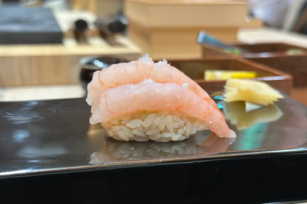 Close-up of a seasonal sashimi sushi piece with translucent shrimp on rice, garnished with flakes. Beside it, a small pile of pickled ginger on a glossy black plate.