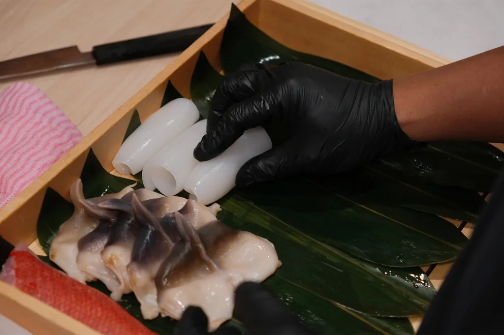 A wooden tray containing fresh sushi ingredients including translucent squid rolls, Hokkigai (surf clam), and kinmedai on green bamboo leaves.
