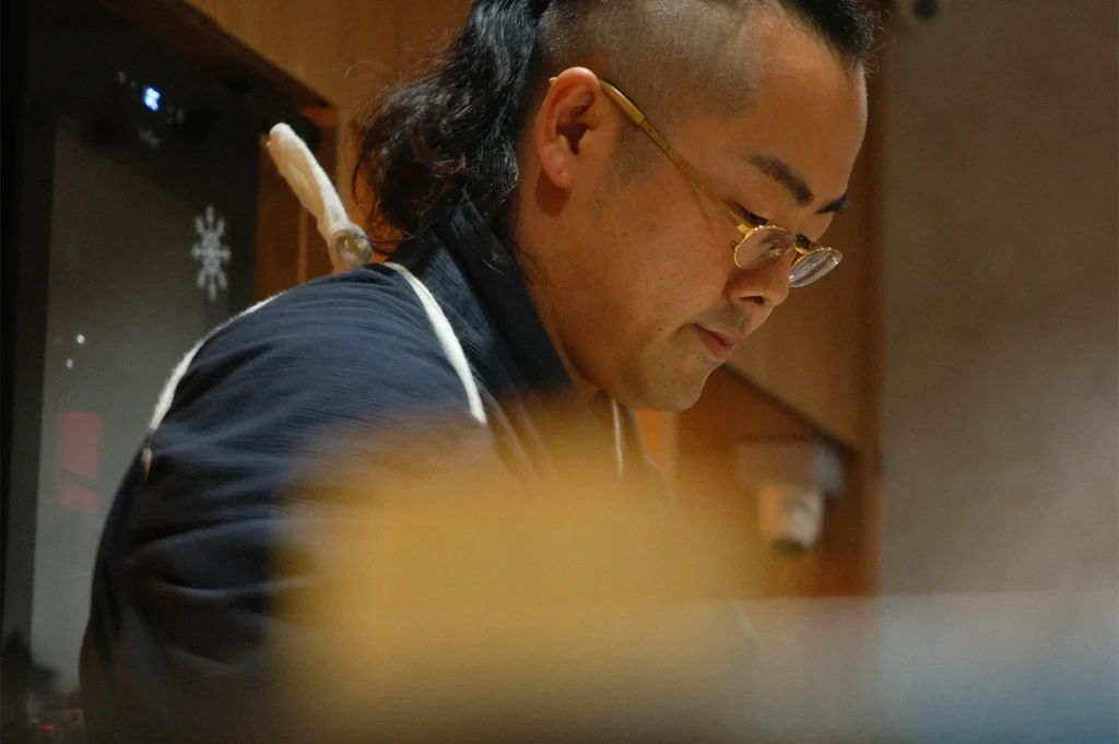 A profile view of Chef Masa with glasses and a traditional uniform, focused on preparing food at a restaurant counter.