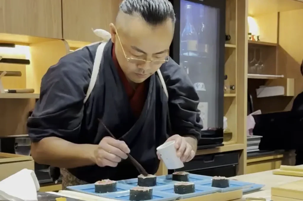 A professional sushi chef with glasses and a traditional dark uniform meticulously garnishing pieces of nigiri sushi on blue ceramic plates.