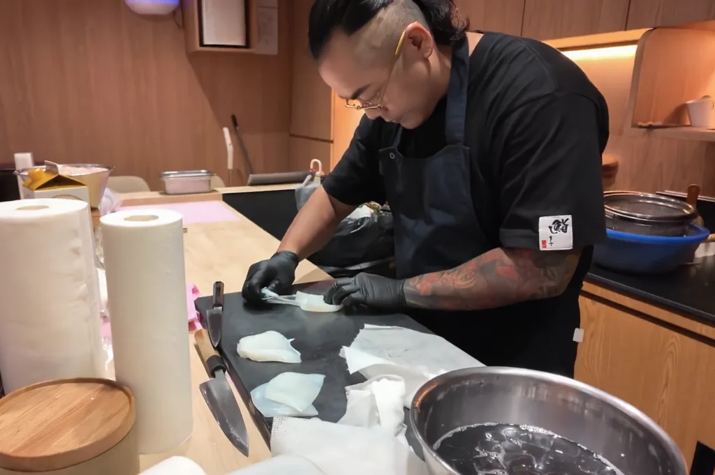 A professional sushi chef with tattoos and glasses meticulously slicing fresh white squid (ika) on a black cutting board in a modern kitchen.