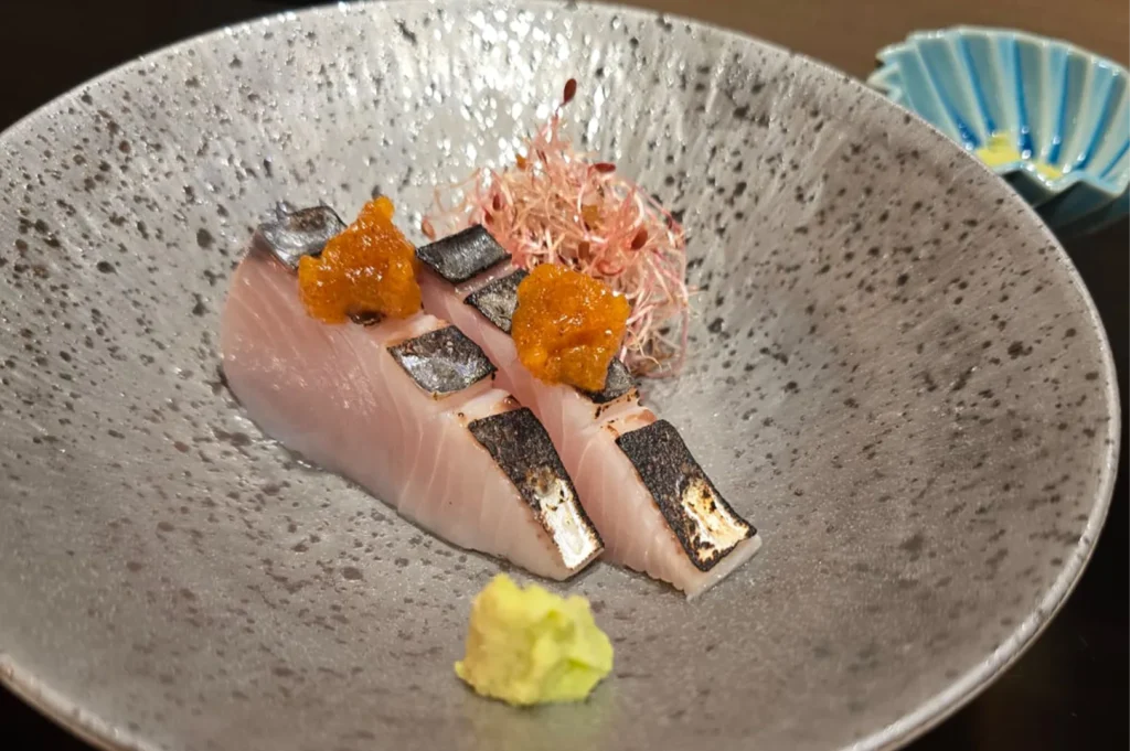 Slices of seared Katsuo (Bonito) sashimi topped with ginger garnish and served with wasabi in a textured grey stone bowl.