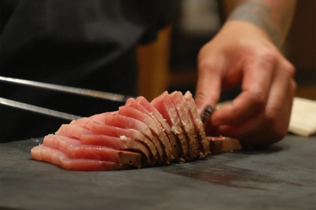 A professional chef slicing a row of seared tuna sashimi on a dark cutting board using precision tongs and a knife.