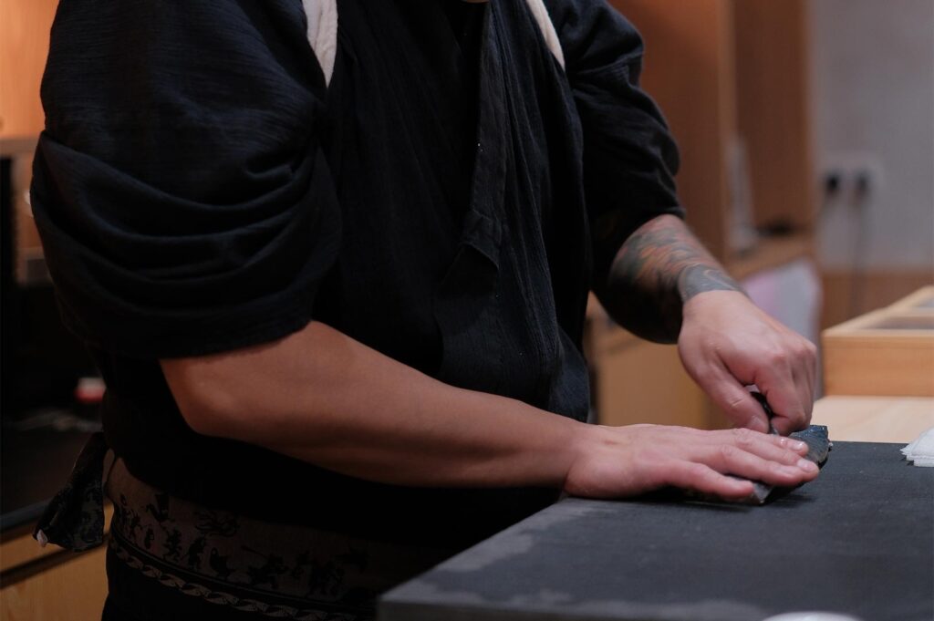Professional sushi chef with traditional attire and tattoos meticulously slicing fresh fish on a workspace.