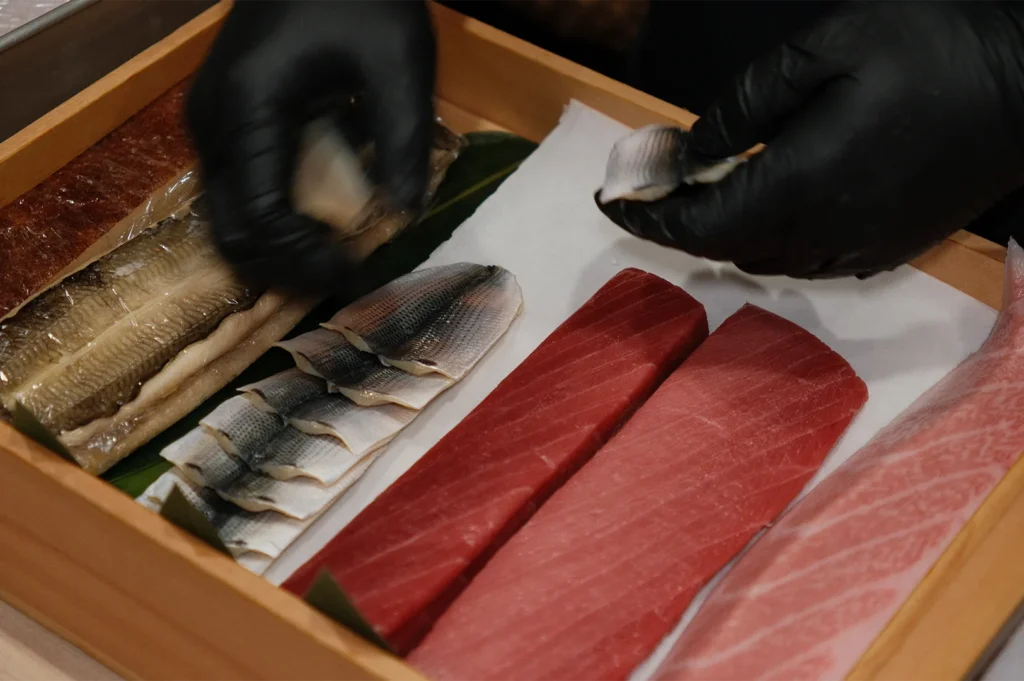 A sushi chef in black gloves preparing various fish on a traditional wooden neta-bako box.