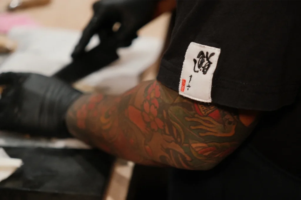 Detailed close-up of a sushi chef with a traditional Japanese tattoo sleeve wearing black gloves while preparing fresh fish.