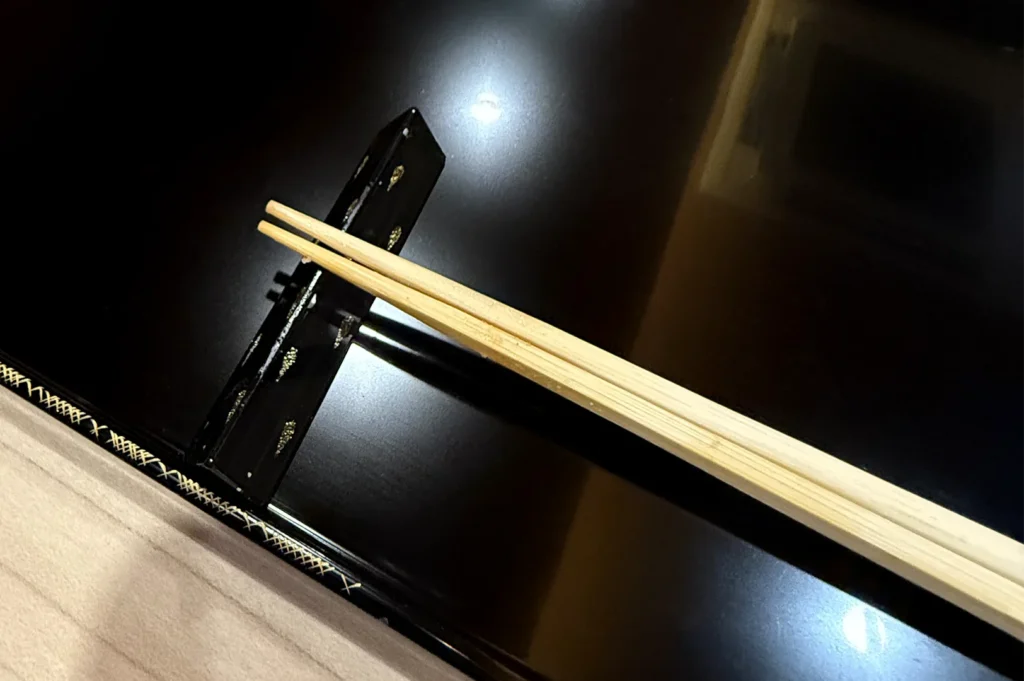 A pair of light wood chopsticks resting on a black lacquer rest over a glossy dark table setting in a fine dining restaurant.