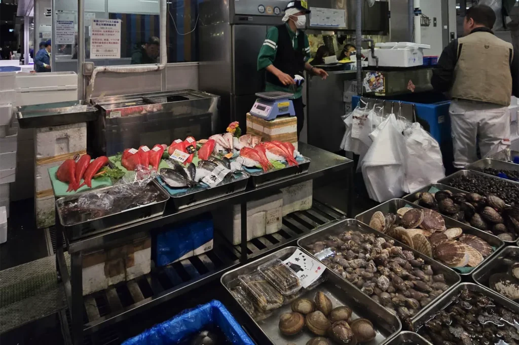 A vibrant display of fresh seafood at a Japanese fish market featuring red snapper, scallops, clams, and various shellfish in metal trays.