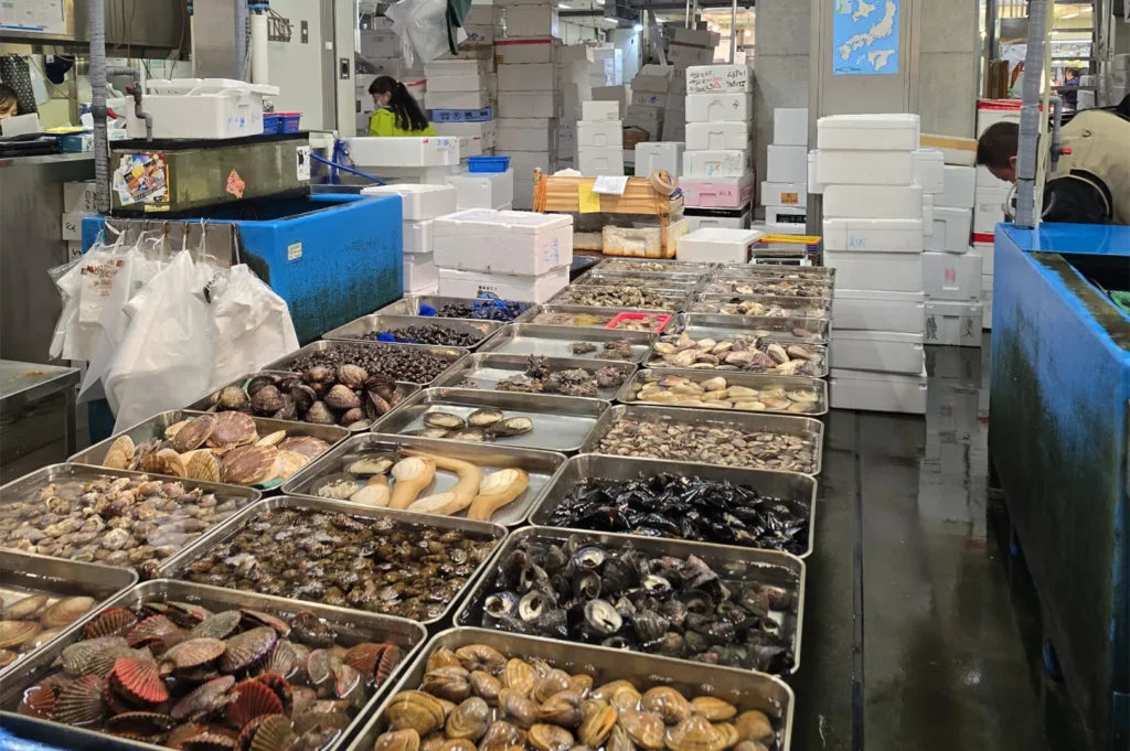 A wide variety of fresh shellfish, including scallops, clams, and mussels, displayed in metal trays at a Japanese fish market.