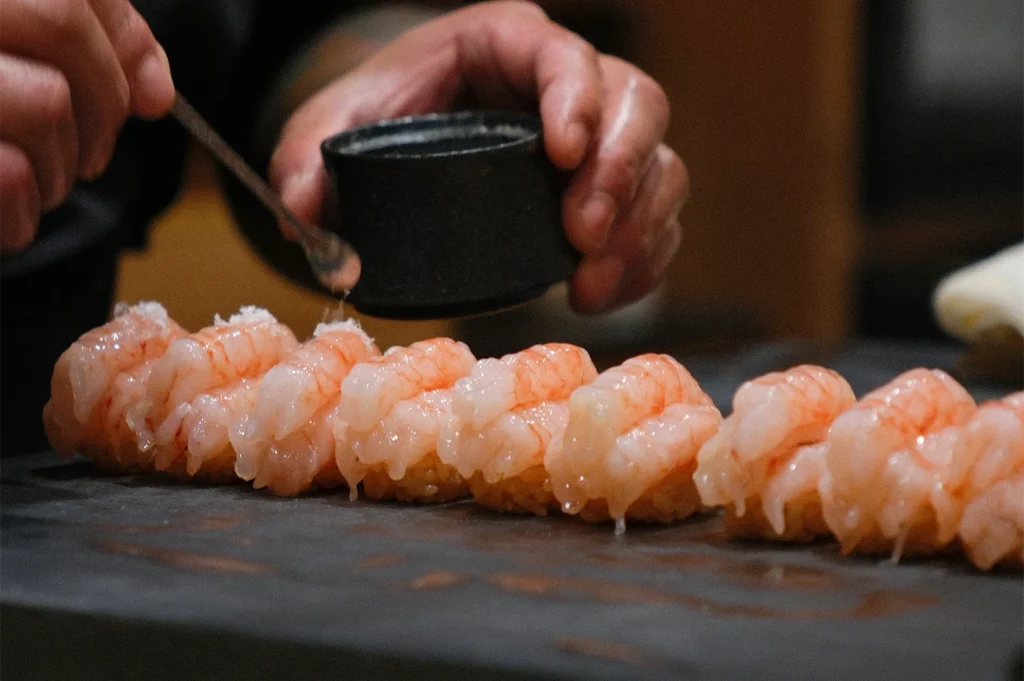 Close-up of a sushi chef meticulously seasoning a row of fresh Ama Ebi (sweet shrimp) nigiri on a black slate serving board.