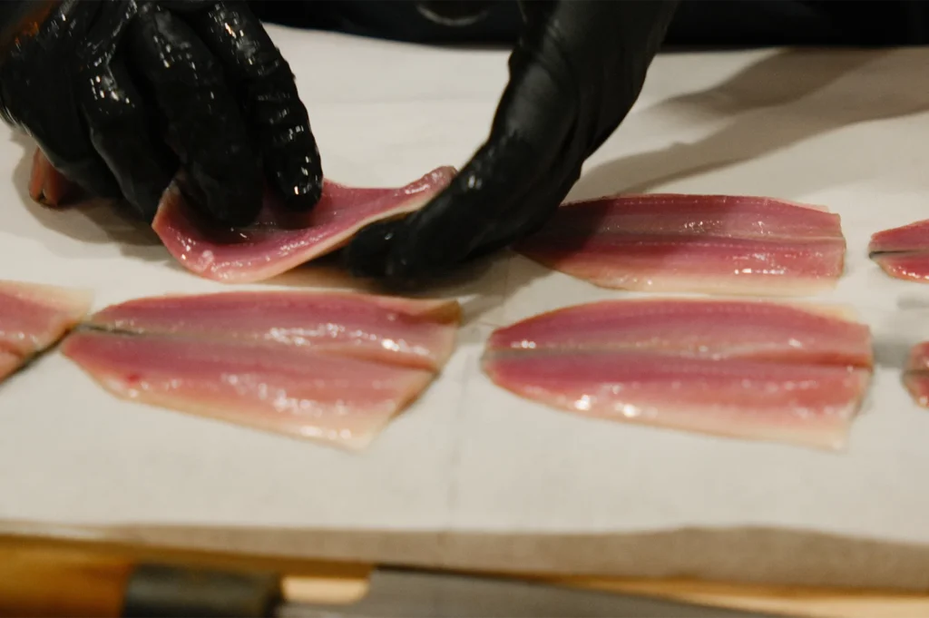 A sushi chef wearing black gloves carefully preparing and slicing fresh pink fish fillets on parchment paper.