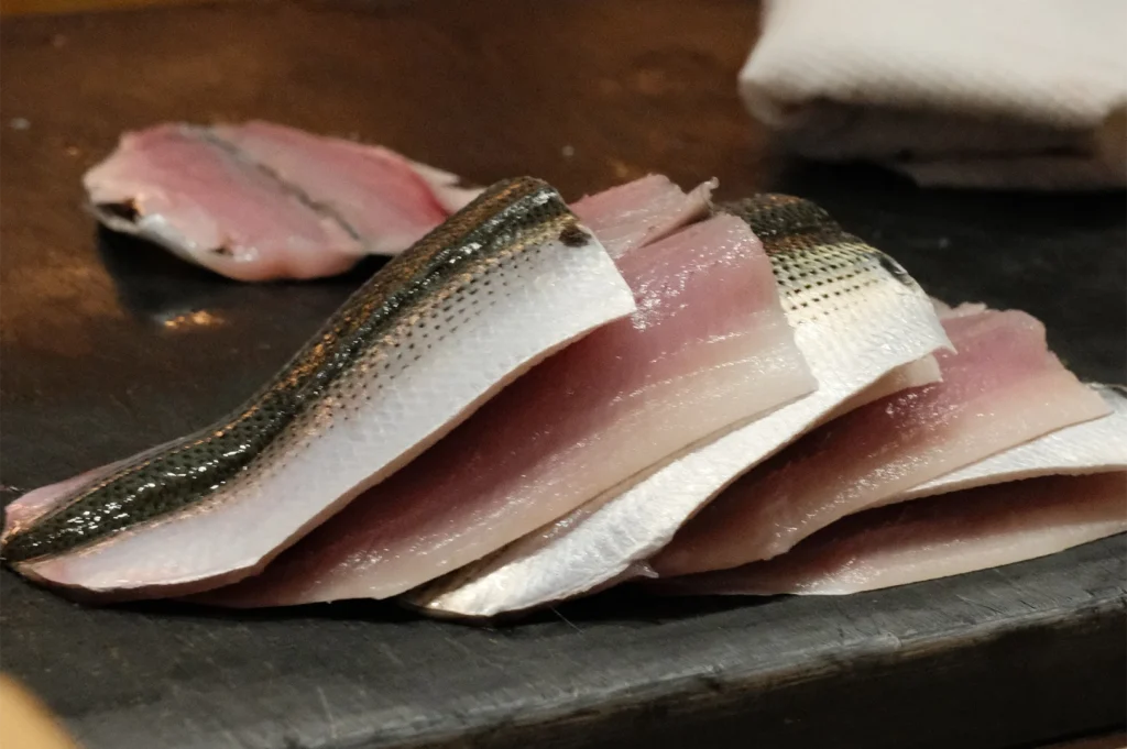 A stack of fresh, salt-cured Kohada (Gizzard Shad) fillets ready for nigiri sushi preparation in a professional kitchen.