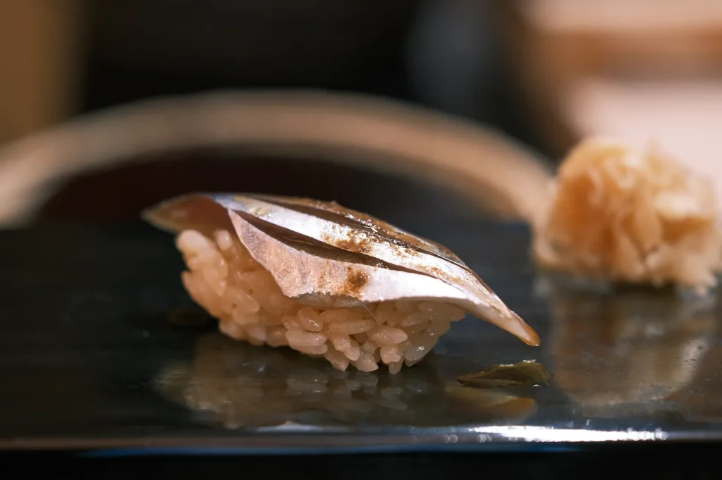 Close-up of a finished Kohada nigiri sushi piece on a black plate with pickled ginger in the blurred background.