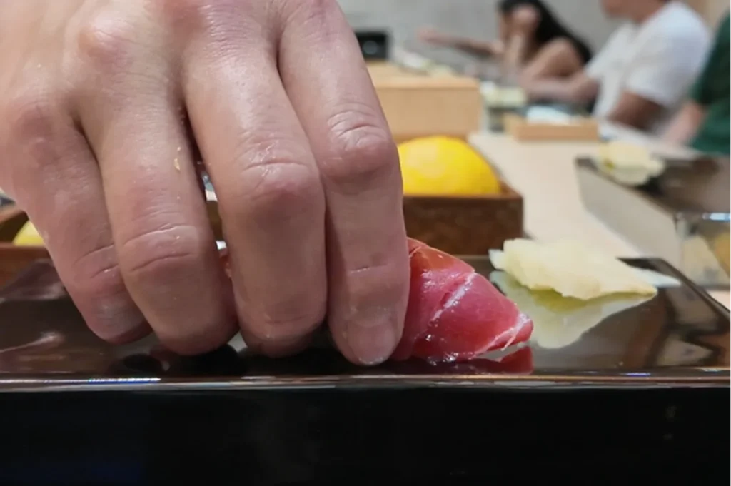 A sushi chef's hand placing a freshly prepared piece of tuna nigiri onto a black ceramic serving plate next to pickled ginger.