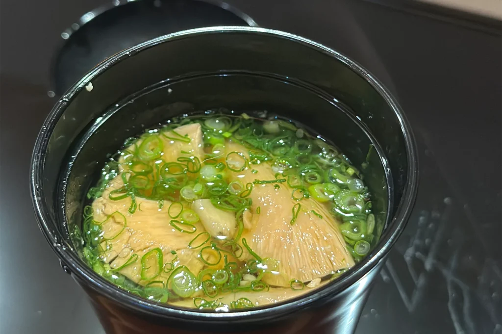 A traditional Japanese clear soup served in a black lacquer bowl, garnished with sliced matsutake mushrooms and fresh green scallions.