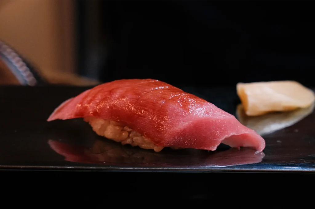 A single piece of premium Maguro (bluefin tuna) nigiri sushi with a deep red color and glossy glaze, served on a black plate with ginger.