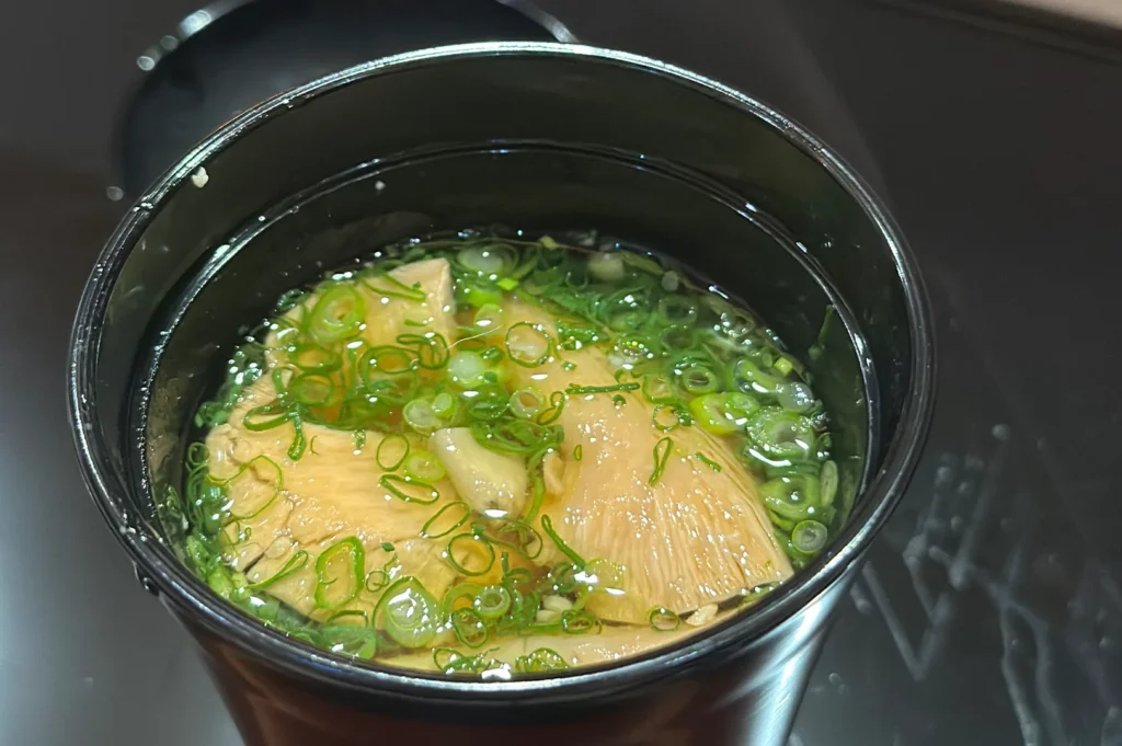 A top-down view of a traditional Japanese clear soup served in a black lacquer bowl, garnished with sliced bamboo shoots and fresh green scallions.