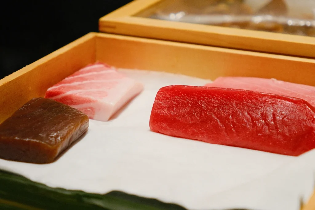 Various cuts of raw tuna, including Otoro and Akami, presented on white paper inside a traditional wooden sushi box.