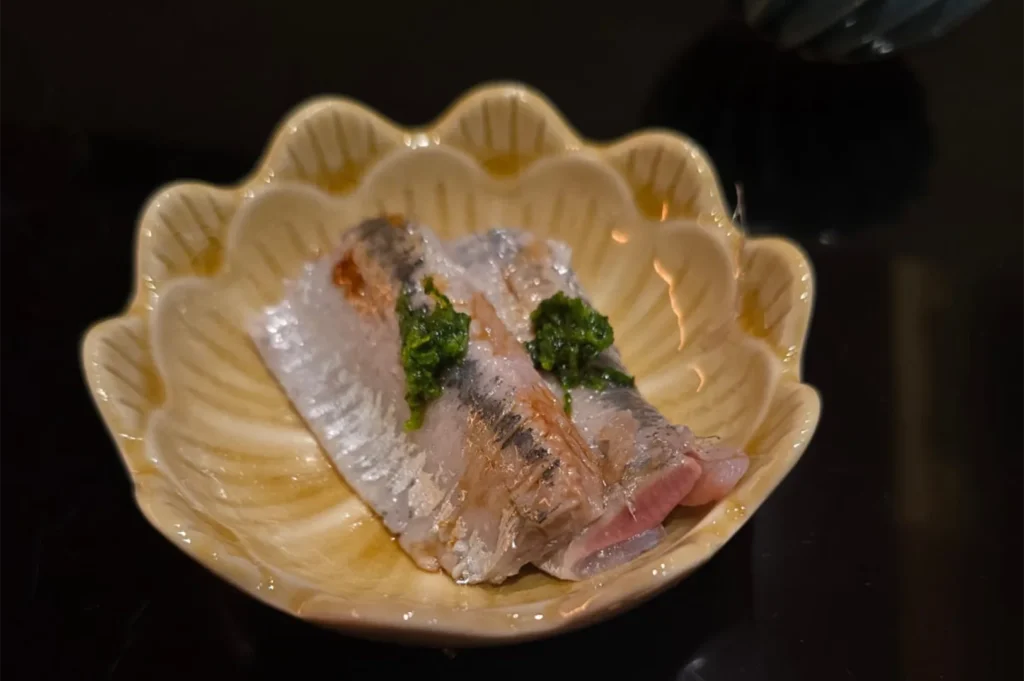 Slices of fresh silver-skin fish sashimi topped with grated ginger and green onions in a decorative scalloped ceramic bowl.