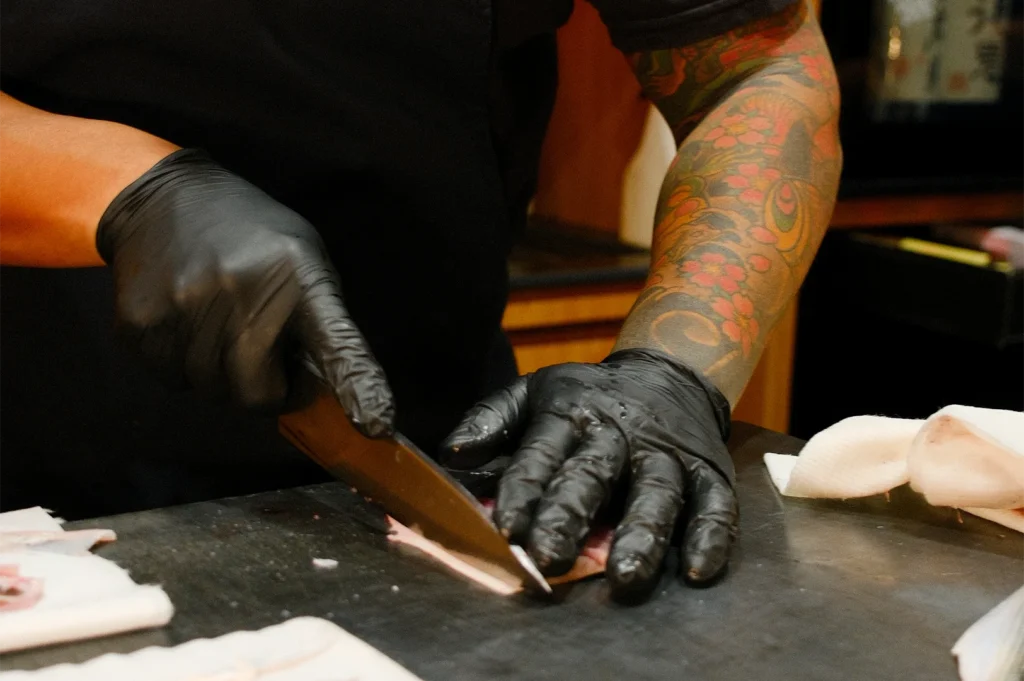 Close-up of a professional sushi chef with a traditional Japanese tattoo sleeve expertly slicing fresh fish using a yanagiba knife.