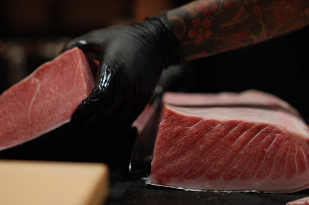 A chef's hand in a black glove lifting a thick, marbled bluefin tuna saku block during the butchery process.