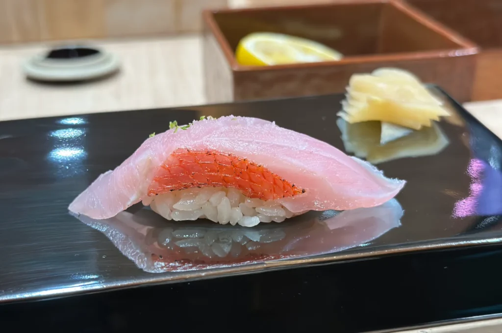 Close-up of a piece of nigiri sushi on a black plate, featuring delicate texture, pink fish atop rice. Pickled ginger and a lemon slice are in the background.