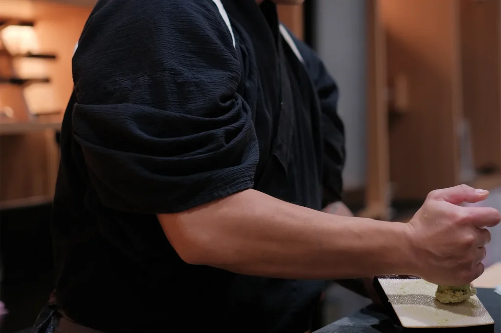 A sushi chef in traditional black attire grating fresh wasabi root on a sharkskin grater (oroshigane).