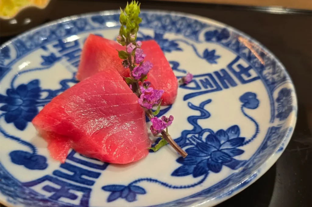 Two thick slices of vibrant red bluefin tuna sashimi garnished with a purple flower sprig on a traditional blue and white patterned ceramic plate.