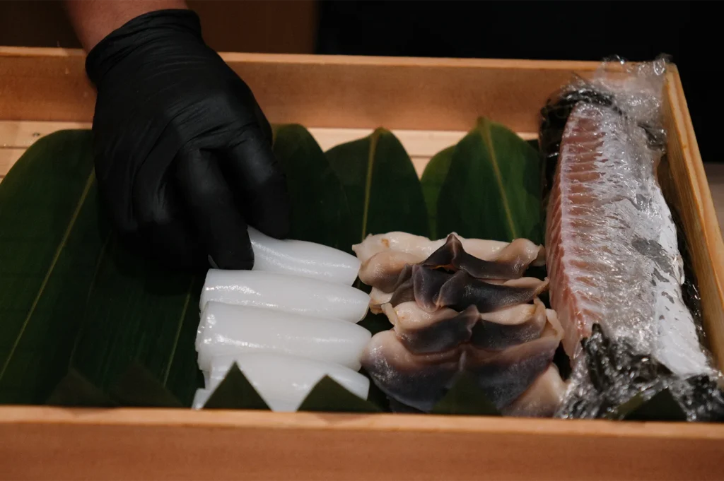 A wooden box containing fresh sushi ingredients including sliced squid, surf clam, and fish skin on green leaves.