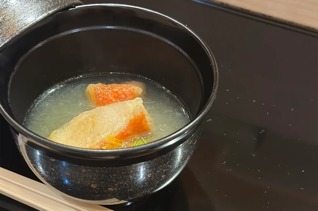 A traditional Japanese clear fish soup served in a black lacquer bowl featuring pieces of cooked white fish and citrus zest.