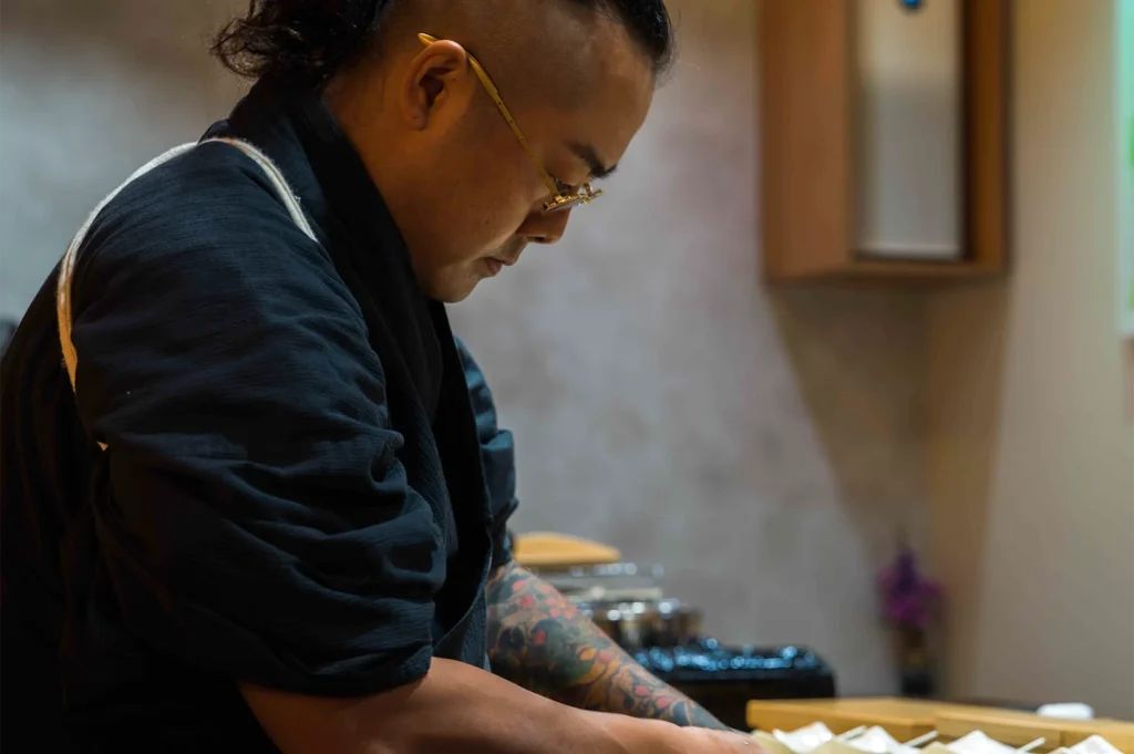 Side profile of a focused professional sushi chef with glasses and a traditional hairstyle working at a wooden sushi counter.