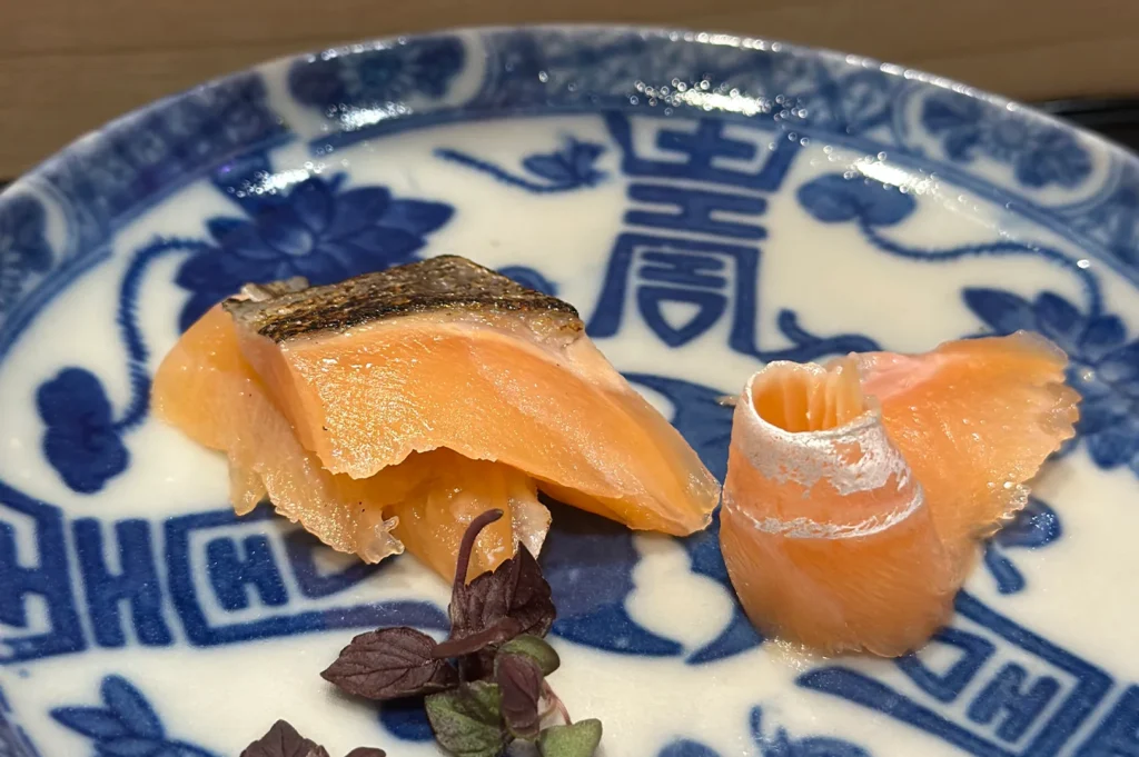 Two pieces of premium seared salmon sashimi, one featuring a silver skin curl, served on a traditional blue and white patterned ceramic Japanese plate.